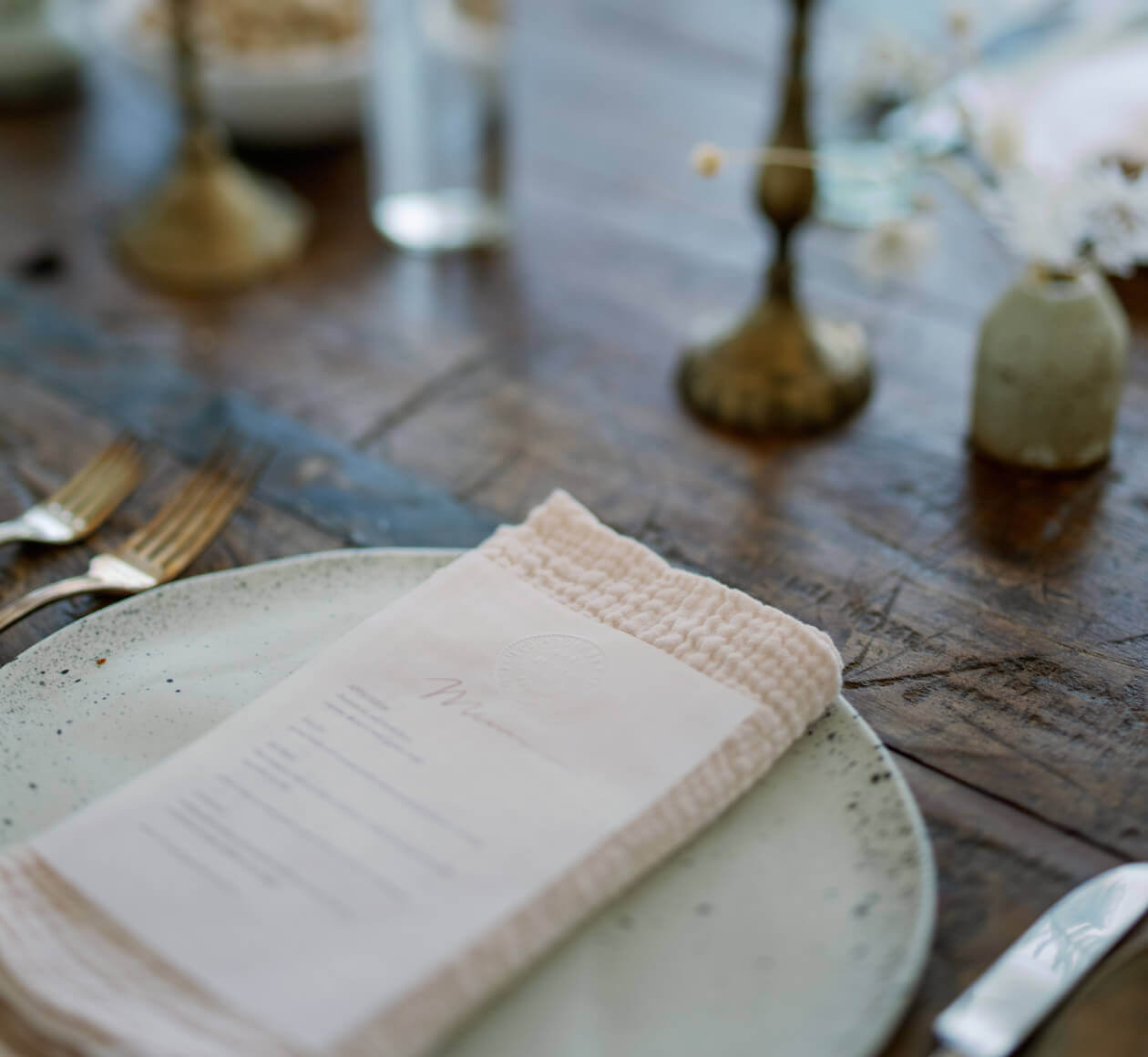 Table setting with a speckled plate, beige napkin, and silverware on a wooden table; soft-focus background with vases and flowers.