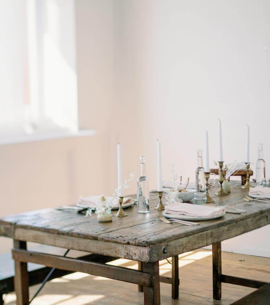 Rustic wooden dining table set for a formal meal with tall white candles, glass bottles, and white napkins in a sunlit room.