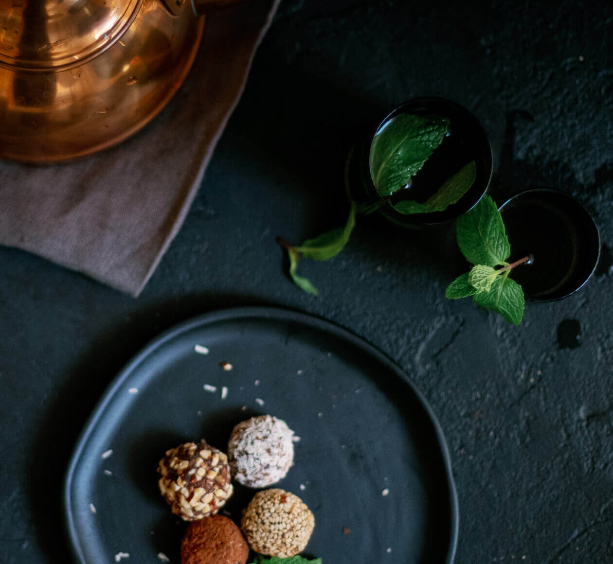 Dark blue plate with four coated truffles on a dark surface, a mint sprig in a glass and a copper kettle in the background.